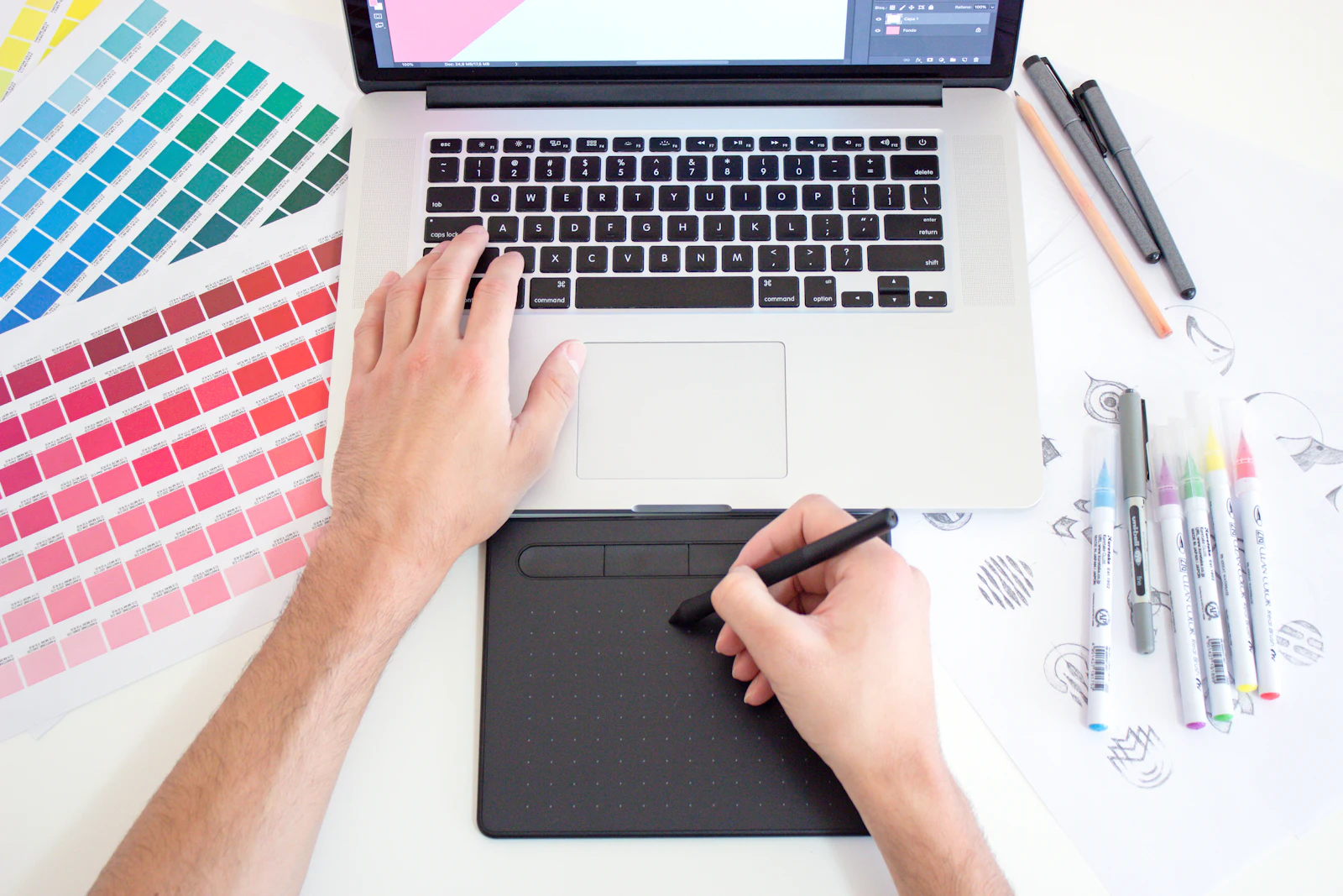 Colour swatch sheets, sketches and a pen tablet beside a laptop, seen from above.