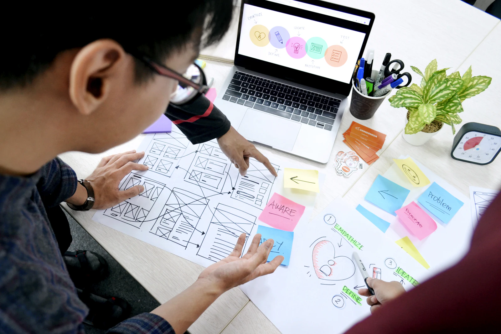Two people planning a website on paper: hand-drawn wireframes, sticky notes and a laptop on the table.